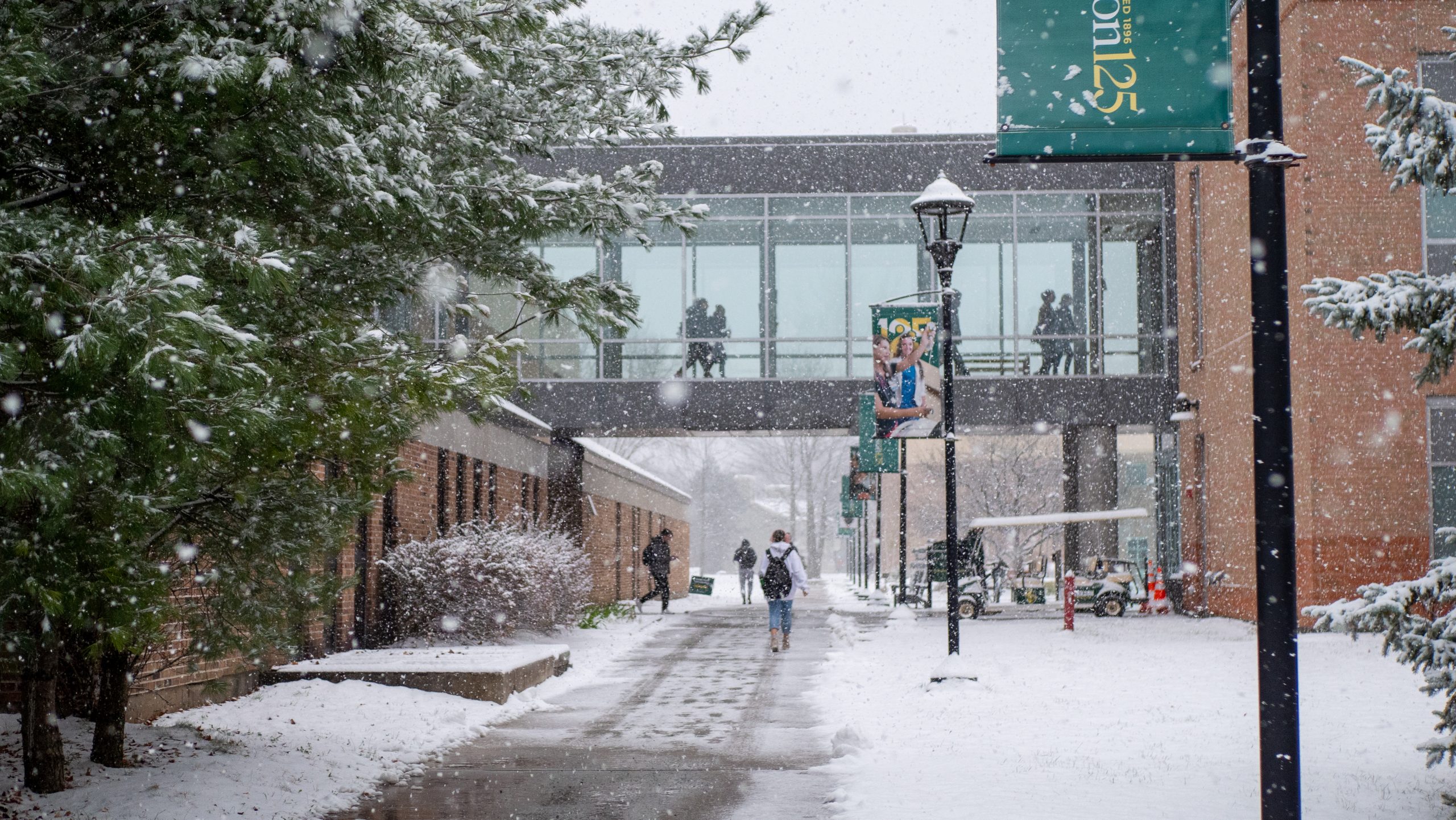 Students walk to class along a paved path on Clarkson's campus, while more students use an elevated indoor pathway to pass between buildings above the outdoor path.