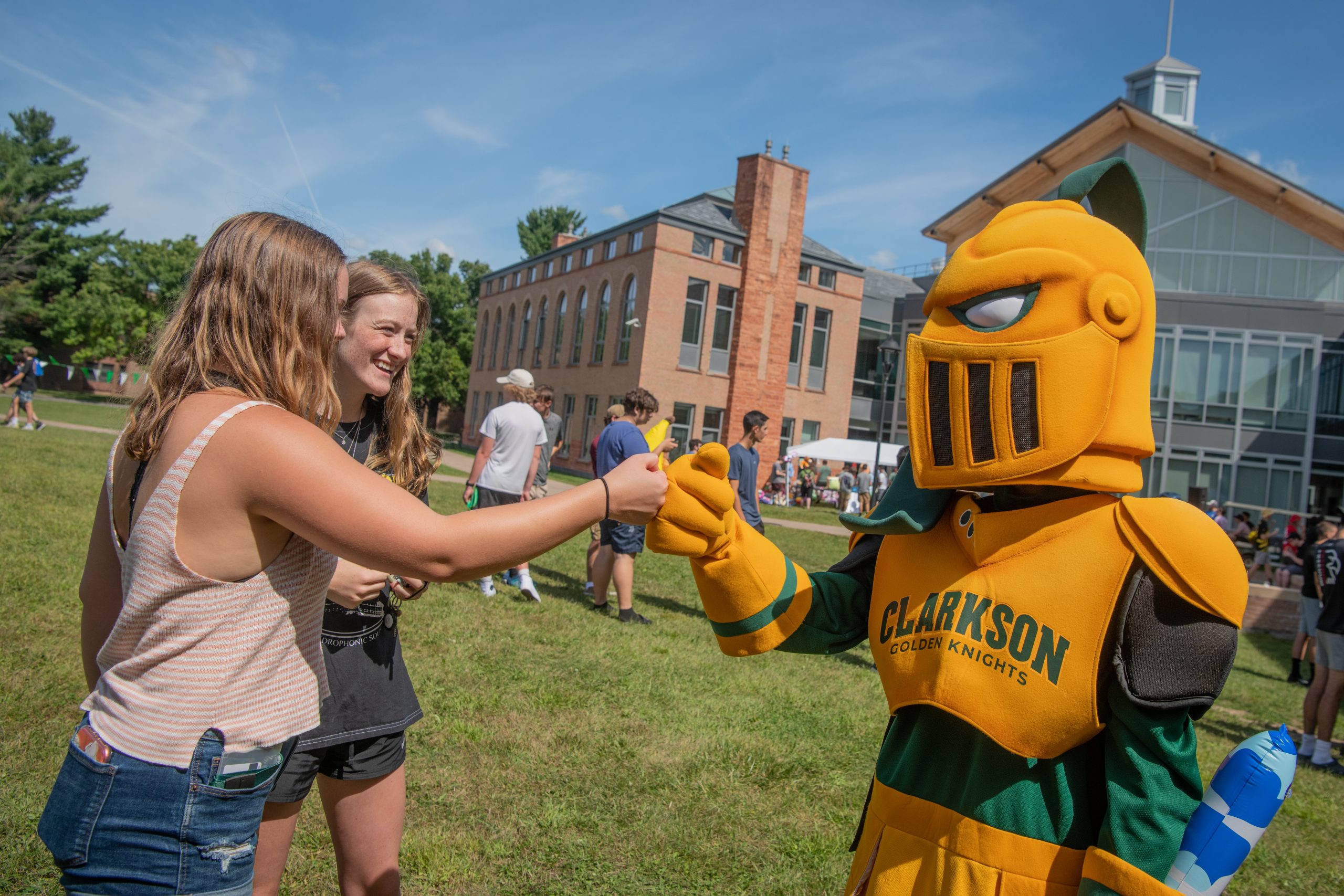 Two Clarkson undergraduate students bump fists with the Golden Knight mascot on the Quad on a summer day.