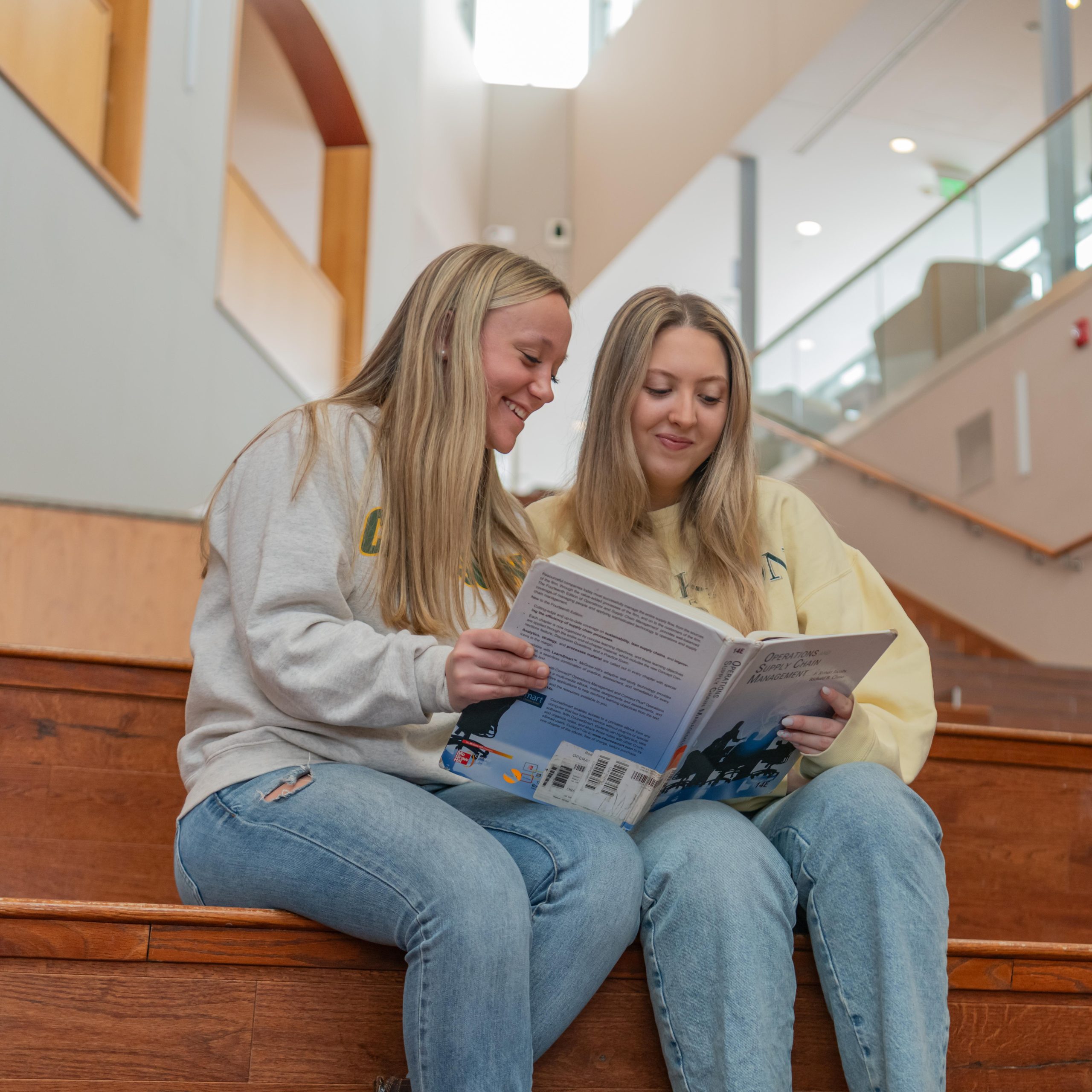 Two women sitting on the steps in the Student Center Forum holding a book