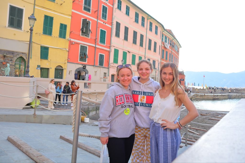 Carly and two other women standing arm in arm against a backdrop of colorful old buildings along a canal