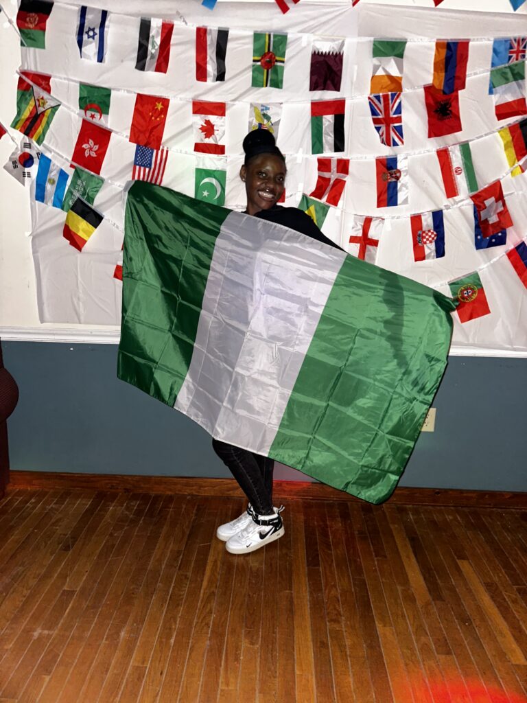 Chidi stands in front of a wall of flags and proudly represents her home country, Nigeria, by showcasing their flag 