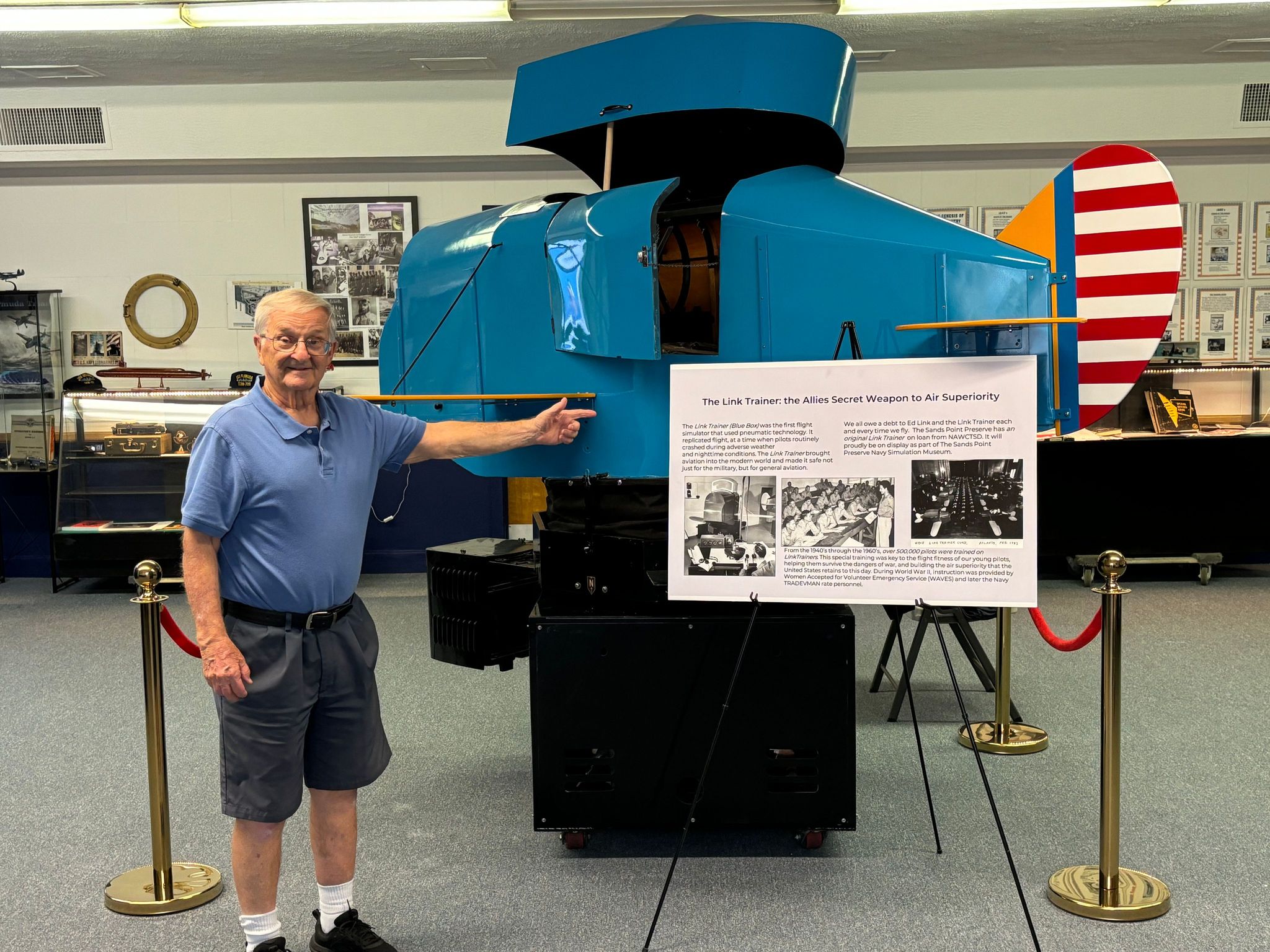 Hank Okraski stands next to and points at a blue Link Trainer antique flight simulation machine.