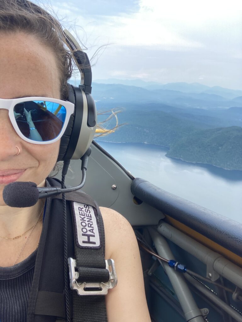 A woman (Heather Woods) wearing a headset with a microphone and white sunglasses takes a selfie while flying in a small aircraft. She has a slight smile, and her reflection in the sunglasses reveals another person, possibly the pilot, in the cockpit. She is secured with a black "Hooker Harness" safety strap. The background showcases a scenic aerial view of a mountainous landscape with a large body of water below. The sky is partly cloudy with soft lighting.
