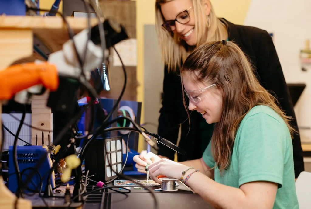 A female student smiles while soldering; a female professor leans over her from the background.
