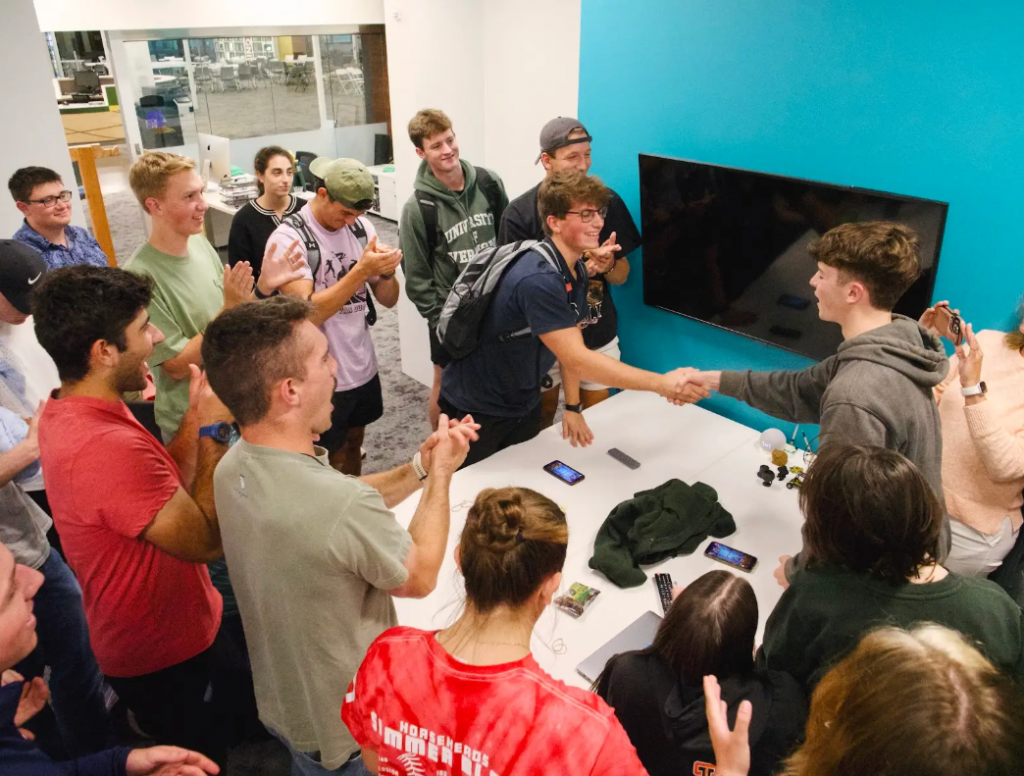A group of students smile and laugh while two shake hands.