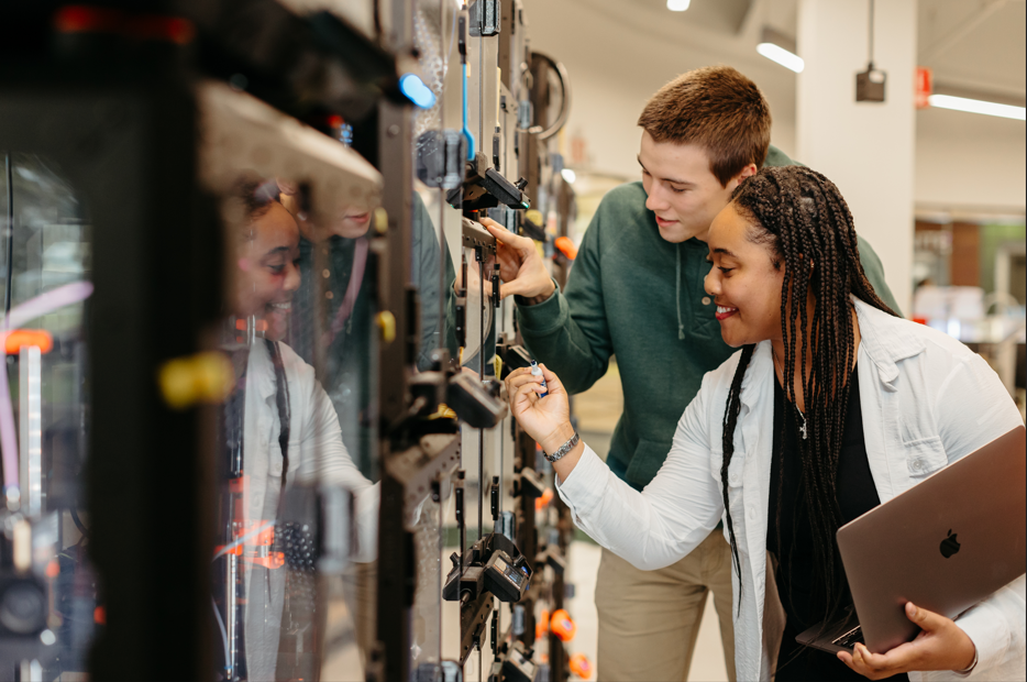 A male and female student smile while examining a 3D printer in the Dorf Makerspace.