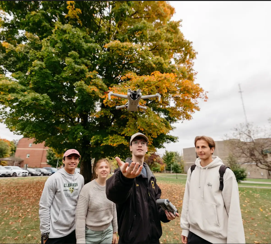A group of four students look up at a flying drone, with one reaching his hand out underneath.