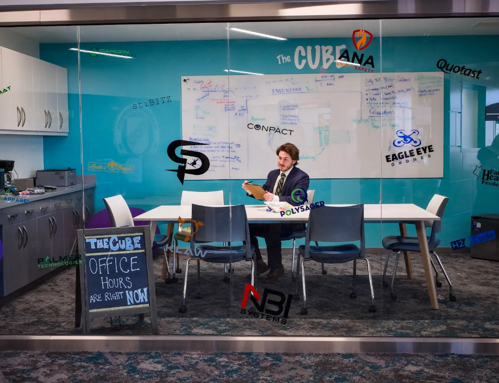 A man sits inside a glass-walled room named the Cube while preparing business plan information using a clipboard.