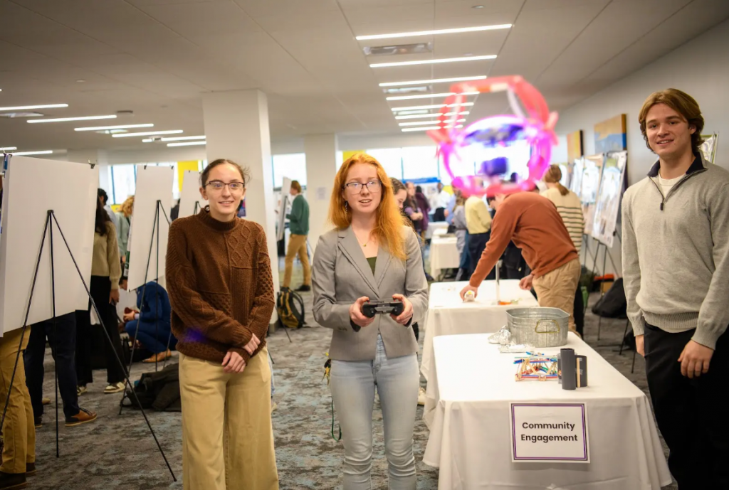 Two students watch while another controls a flying drone featuring flashing lights.

