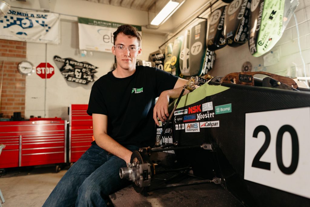 Clarkson University mechanical engineering student poses with a Formula SAE car inside the SPEED Team garage, surrounded by past car panels and tool benches.