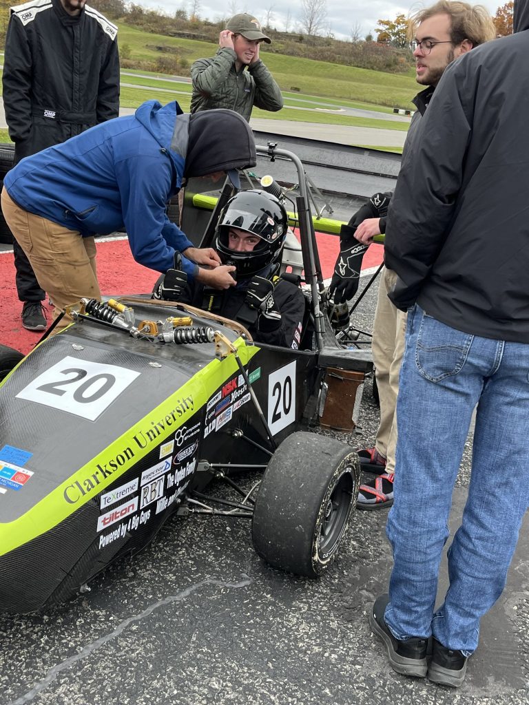 Clarkson University mechanical engineering student prepares for a Formula SAE test drive with teammates assisting trackside.