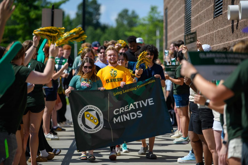 Students from Clarkson University’s Creative Minds Living Learning Community march in a welcome parade, holding a banner and waving pom-poms while being cheered on by upperclassmen and volunteers.