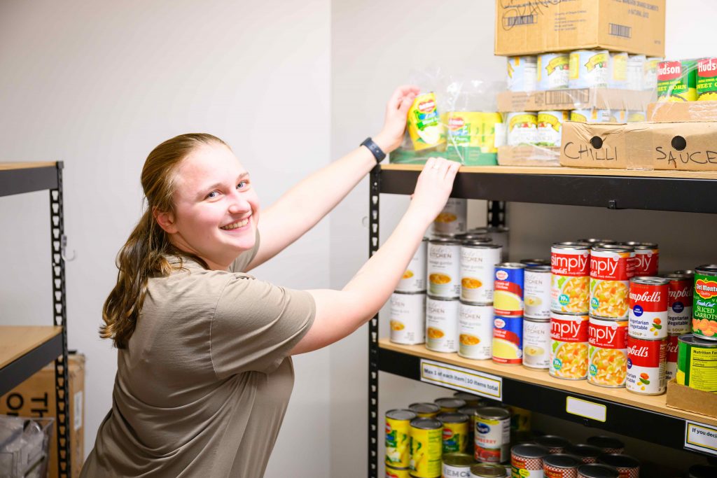 Student smiling while organizing canned goods on a shelf inside the Clarkson University Food Pantry.