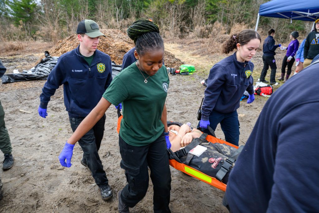 Clarkson University students participate in an EMS training exercise, carrying a mock patient on a stretcher during a simulated mass casualty incident outdoors.