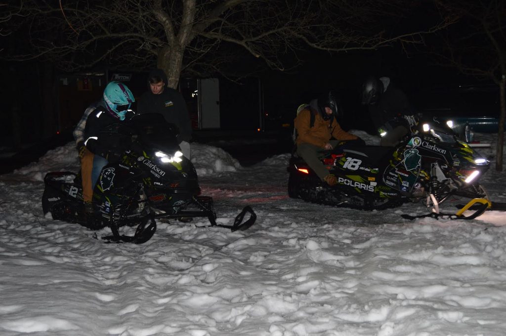 Two Clarkson students prepare for a nighttime test ride, seated on snowmobiles with headlights glowing in the snow.