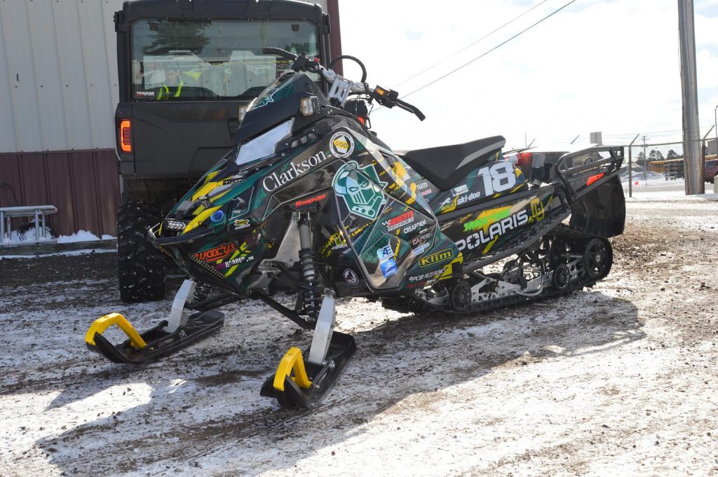 The Clarkson SAE Clean Snowmobile, covered in sponsor logos, is parked outside the shop during winter testing.