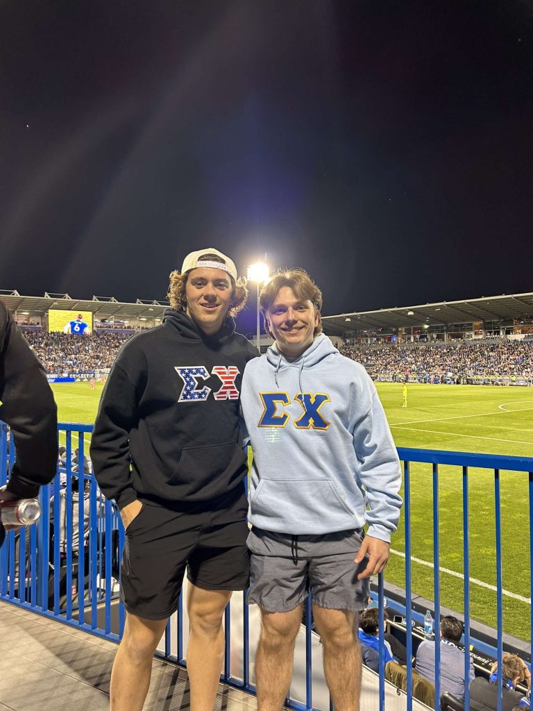 Two young men in hoodies with greek lettering pose for a photo with a crowded soccer match in the background.