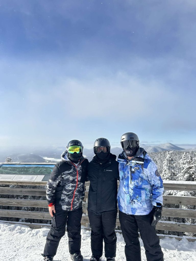 Three young men head-to-toe in ski gear pose for a photo on top of a mountain on a partly cloudy day