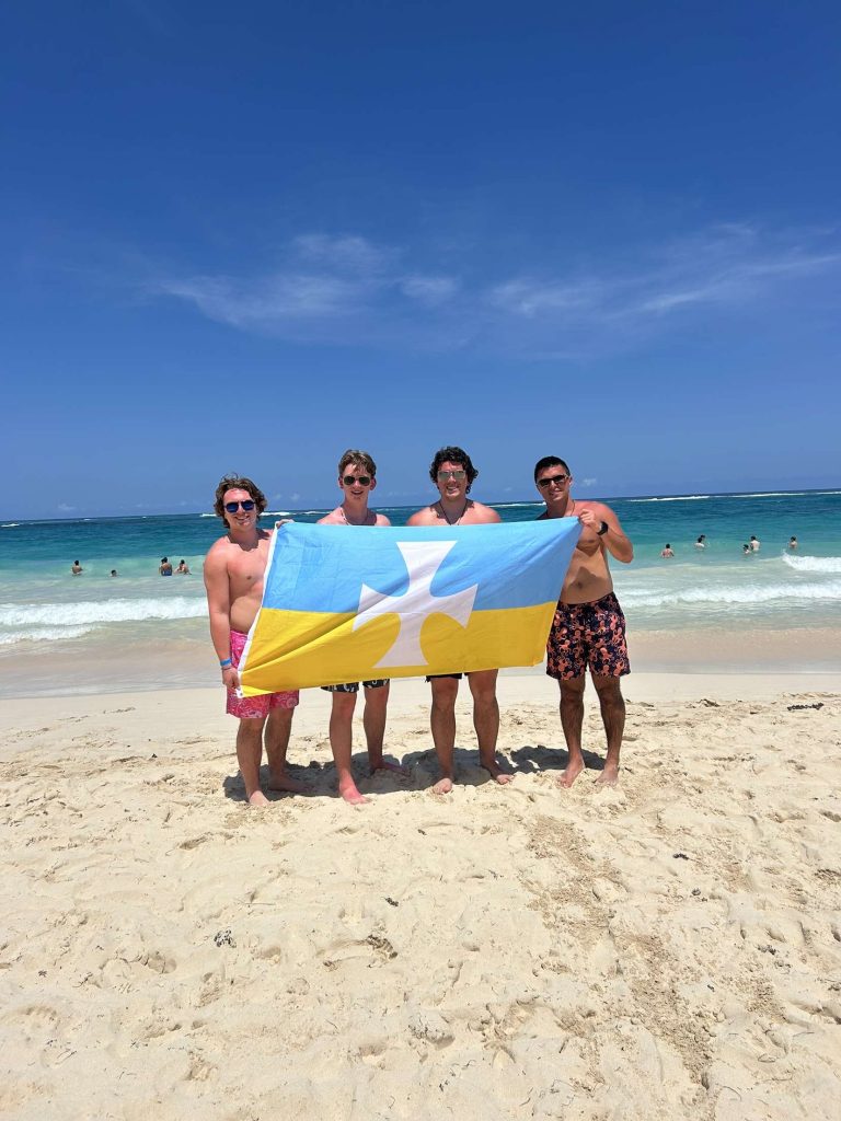 Four young men pose on a beach displaying a blue and yellow Sigma Chi flag.