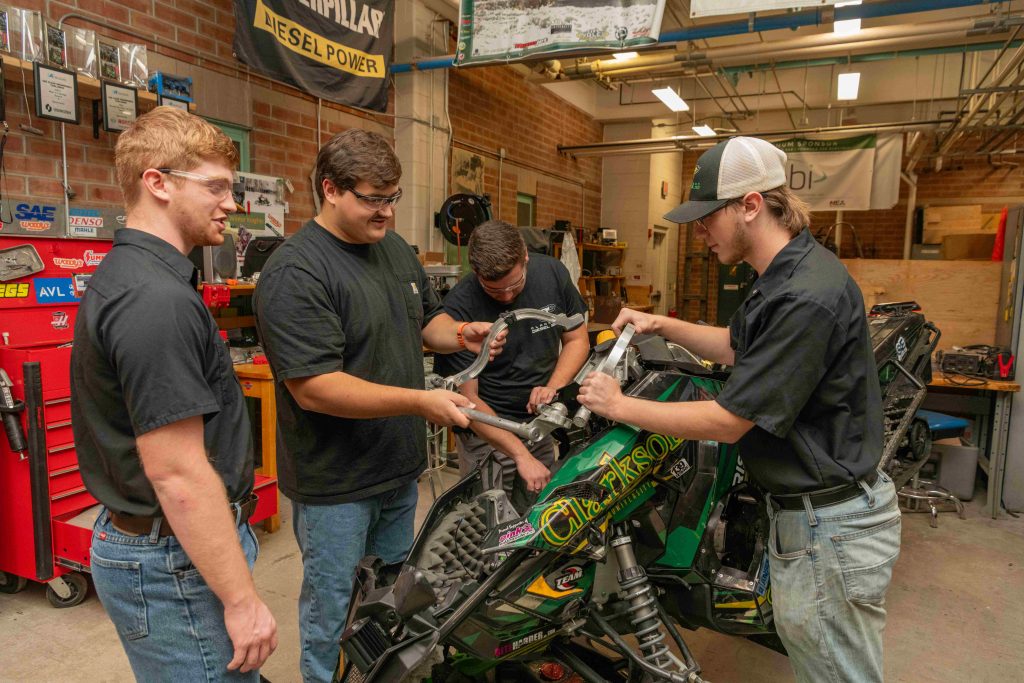 A group of Clarkson SPEED team members collaborates in the shop, working together to assemble a snowmobile chassis.