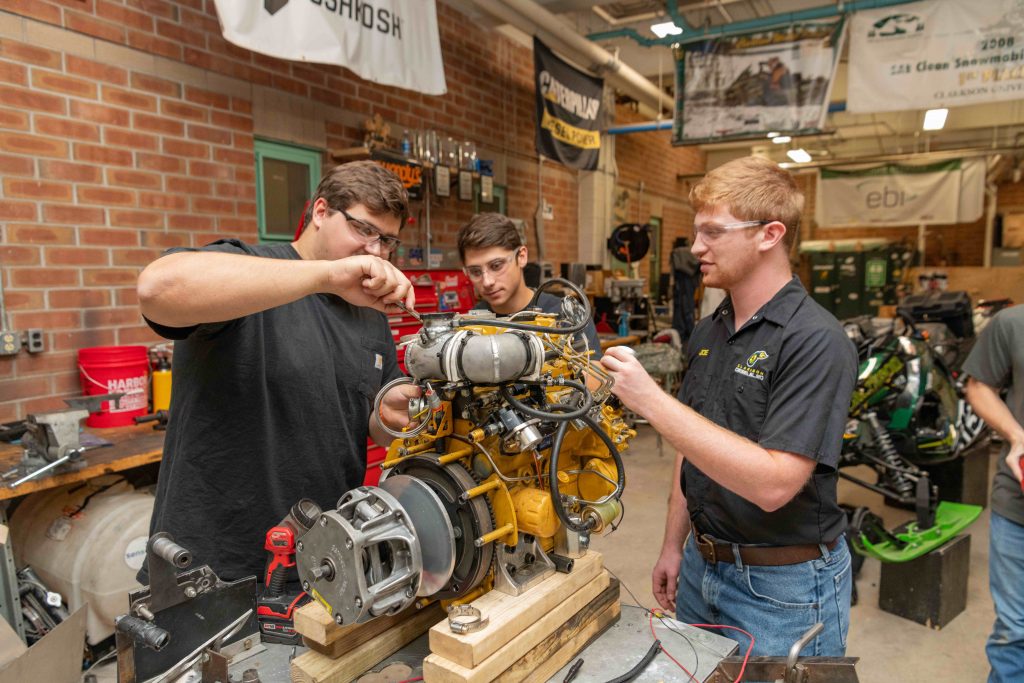 Clarkson students examine and adjust a yellow engine in the SPEED machine shop, surrounded by tools and equipment.