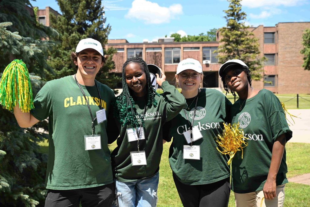 Four students from Clarkson University sport Clarkson gear and nameteags, standing in front of a student housing building on campus.