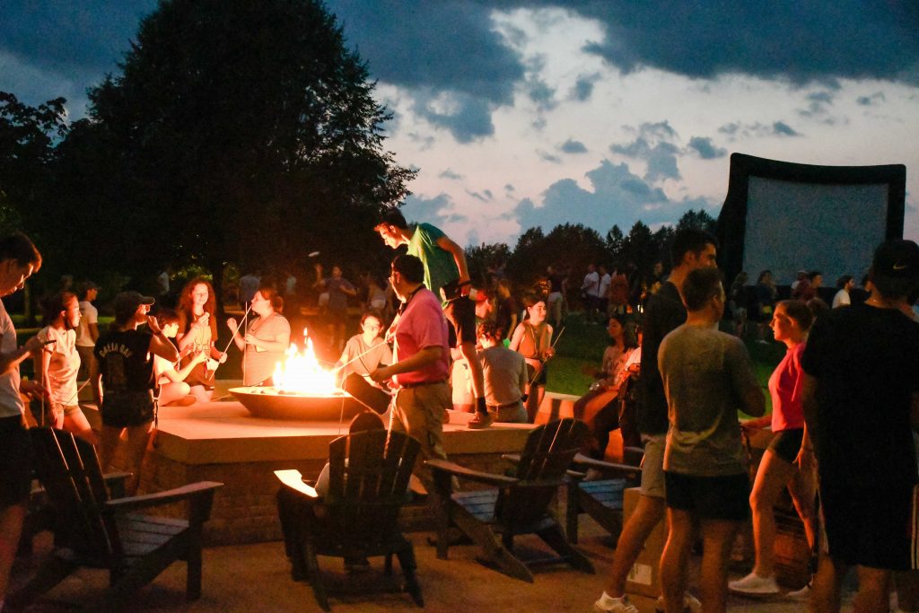 Clarkson students gathered around an outdoor firepit at night, roasting marshmallows and enjoying a campus social event.