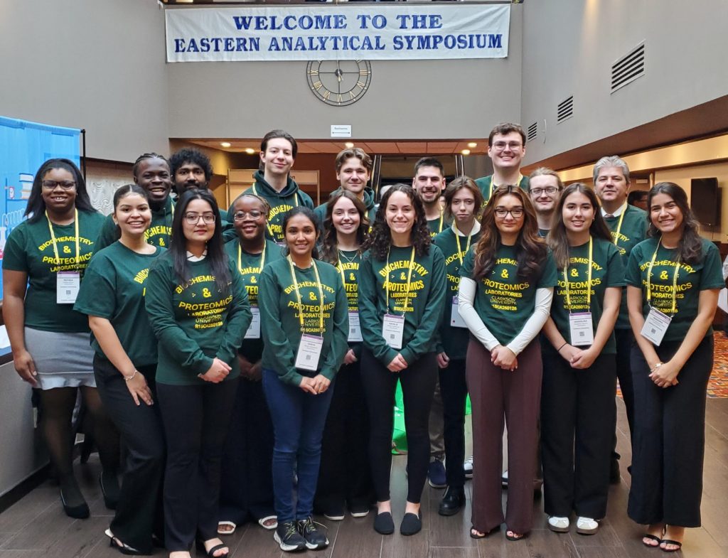 Group of students and faculty wearing matching green ‘Biochemistry & Proteomics Laboratories’ shirts, posing together beneath a banner reading ‘Welcome to the Eastern Analytical Symposium.