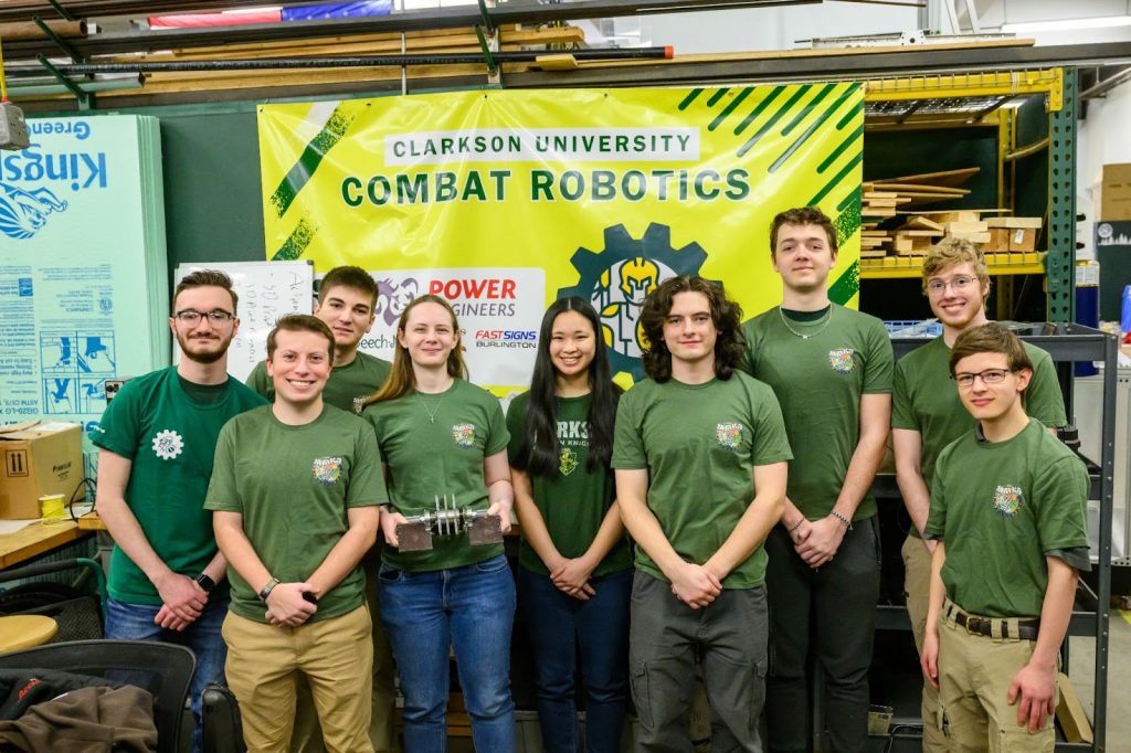 A group of students stand together in front of a banner reading “Clarkson University Combat Robotics”.
