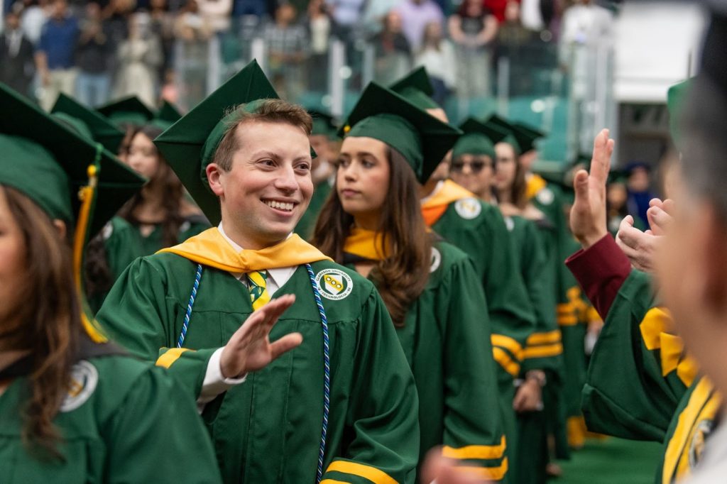 A group of students walk in a line, wearing graduation regalia.
