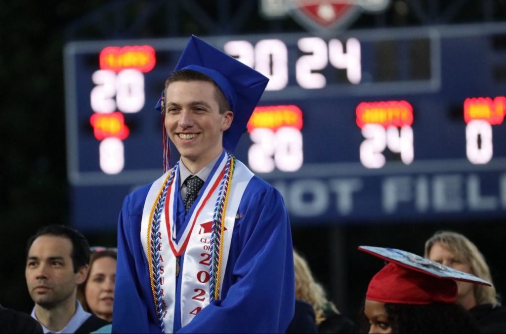 A male student smiles while standing, wearing graduation regalia.
