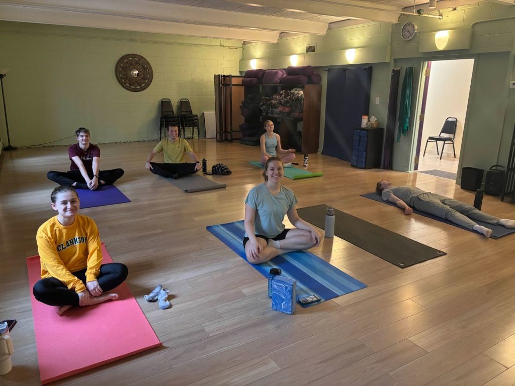 A group of students demonstrate various yoga poses while sitting on yoga mats.
