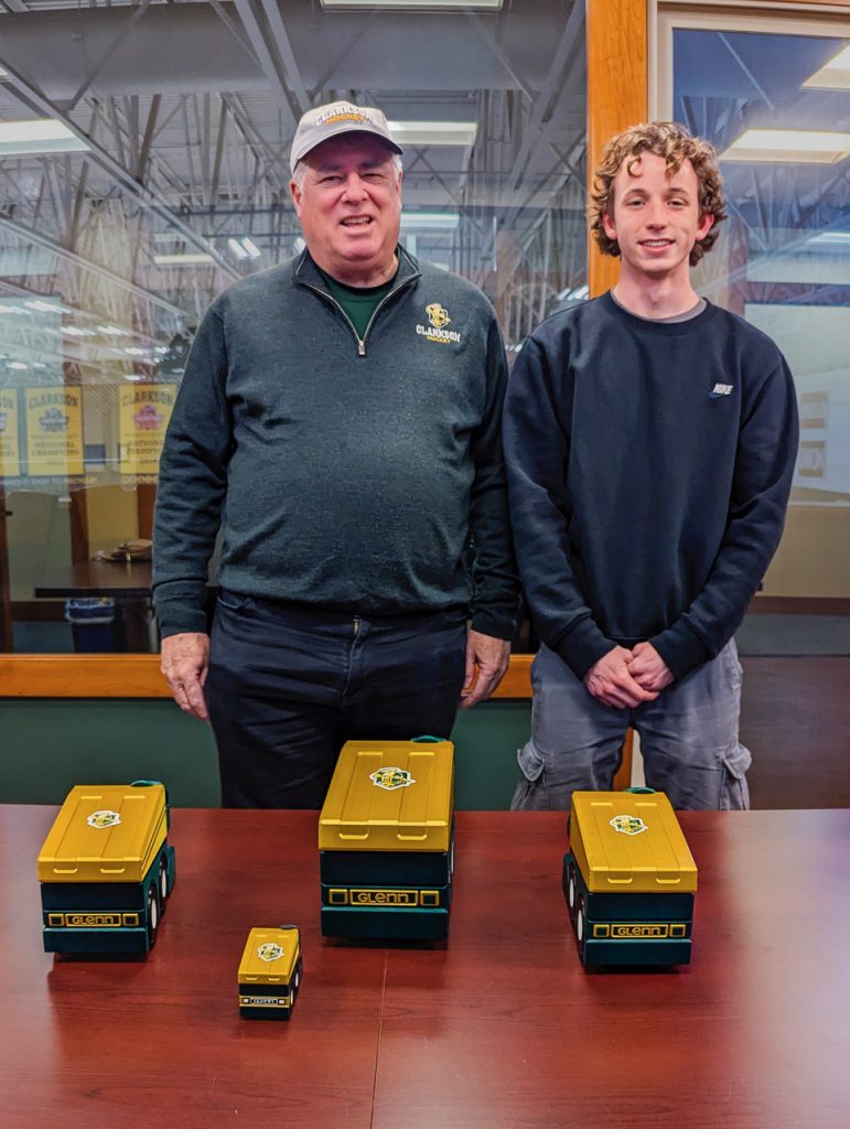This is an image of David Glenn and Daniel Galko. They are smiling for the photo and standing in front of a table. On the table are four replica ice resurfacing machines that are about six inches in height.