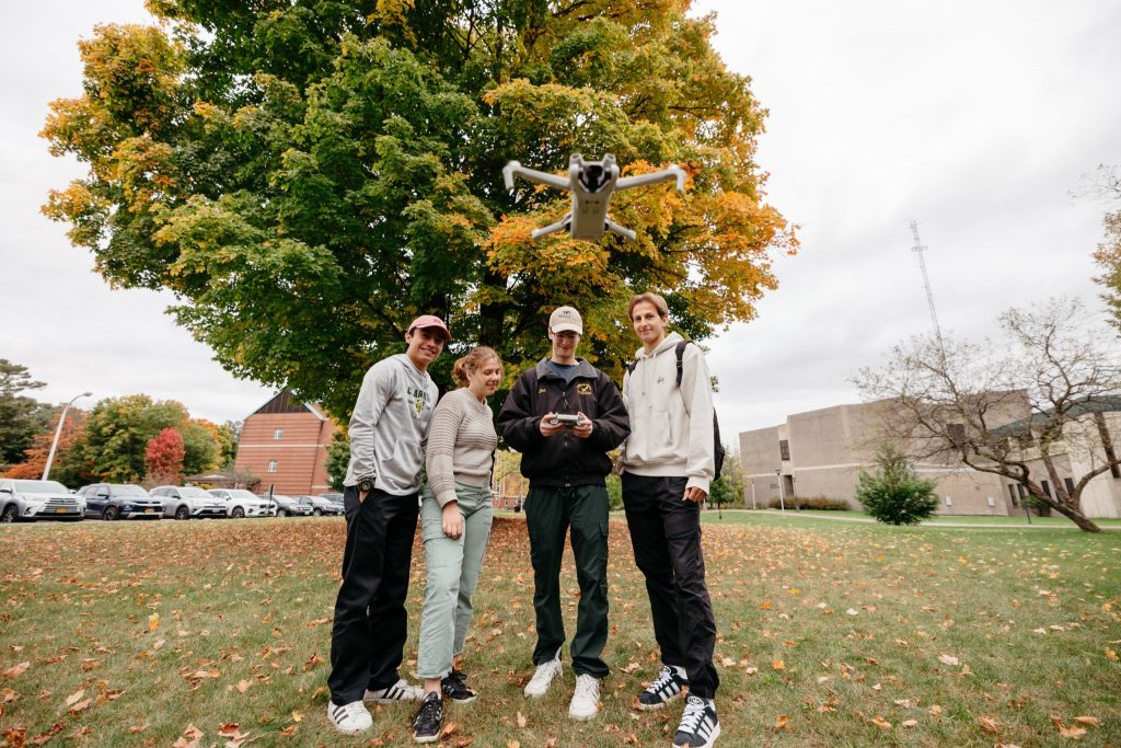 Four Clarkson University students stand on a grassy campus lawn covered in autumn leaves, operating a small white drone. One student holds the remote controller while the others look on. A large tree with changing green and yellow leaves serves as the backdrop, with campus buildings and parked cars visible in the distance.
