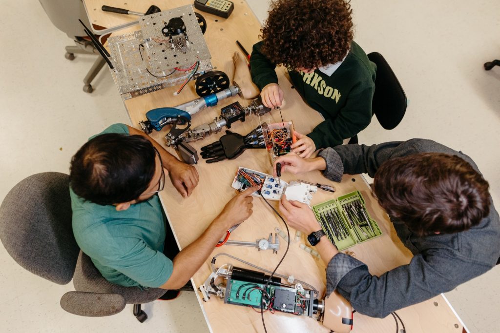 Three Clarkson University students collaborate at a wooden workbench, assembling complex robotic components and prosthetic limbs. The table is filled with tools, including a drill bit set, calipers, and exposed circuit boards, as the students use screwdrivers to fine-tune the mechanical assemblies.
