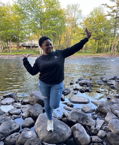This is an image of a young college student who is standing in front of a rocky stream, which is located on a hiking trail. The student is posing for the camera with their arms out. They are smiling for the camera.