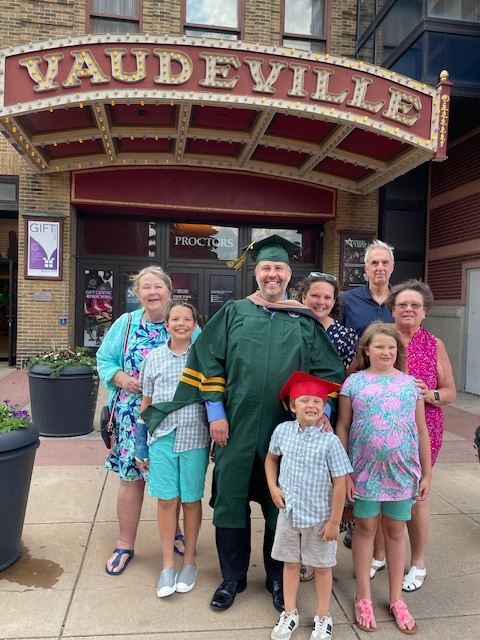 This is an image of a Clarkson University graduate standing in his cap and gown alongside several of his family members in front of a theatre. They are all smiling and happy.