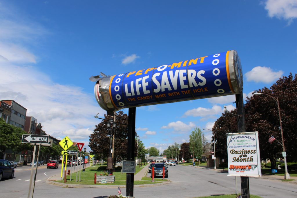 A large, horizontal monument of a Pep-O-Mint Life Savers candy roll, complete with torn silver foil wrapping on the left end, mounted high on black support posts. The sign reads "Pep-O-Mint Life Savers: The Candy Mint with the Hole" in bold white and yellow lettering against a blue background. The monument sits in a grassy median dividing a town street lined with trees, cars, and traffic signs. In the foreground, a smaller white sign reads "Gouverneur Chamber of Commerce Business of the Month."