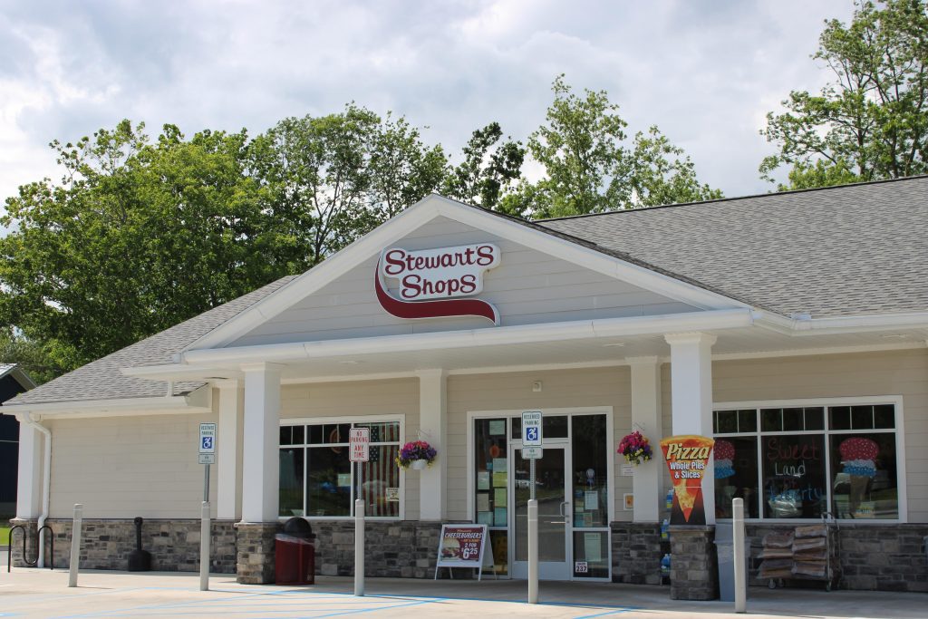 Exterior view of a Stewart's Shops storefront under a partly cloudy sky with green trees in the background. The building has light beige siding, a gray shingled gabled roof, and white supporting pillars resting on stone bases. The prominent "Stewart's Shops" logo is mounted above the main entrance. Windows display advertisements for pizza, cheeseburgers, and a "Sweet Land of Liberty" sign, alongside hanging flower baskets and an American flag visible inside.